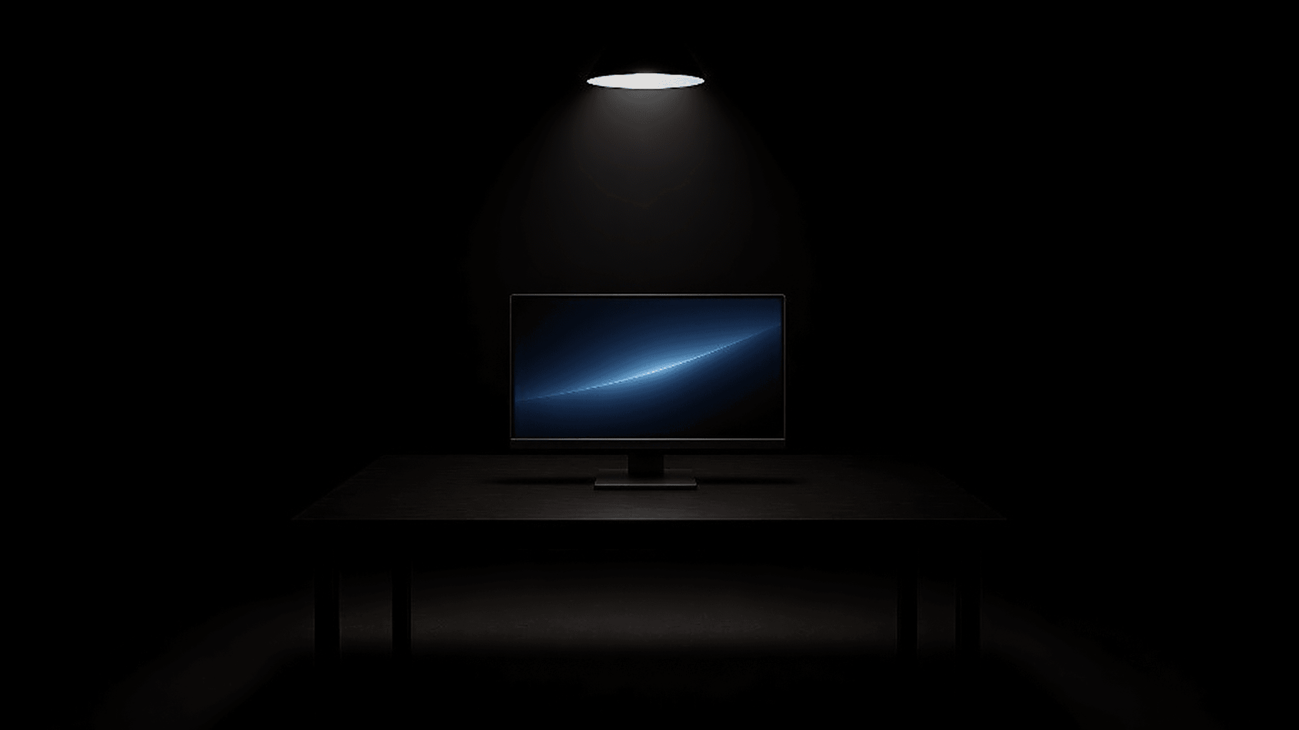 A monitor on a desk in a dark room, lit by a single overhead lamp